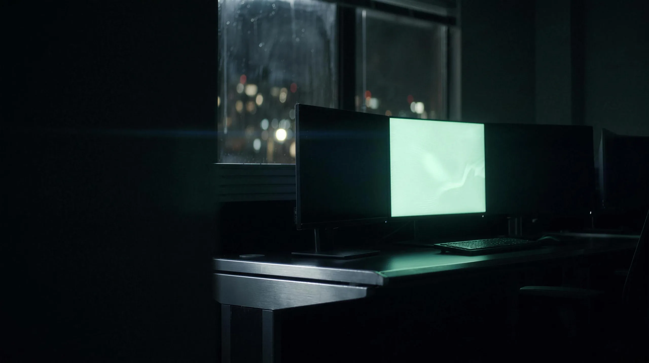 An office at night — monitors glowing mint beside a rain-streaked window